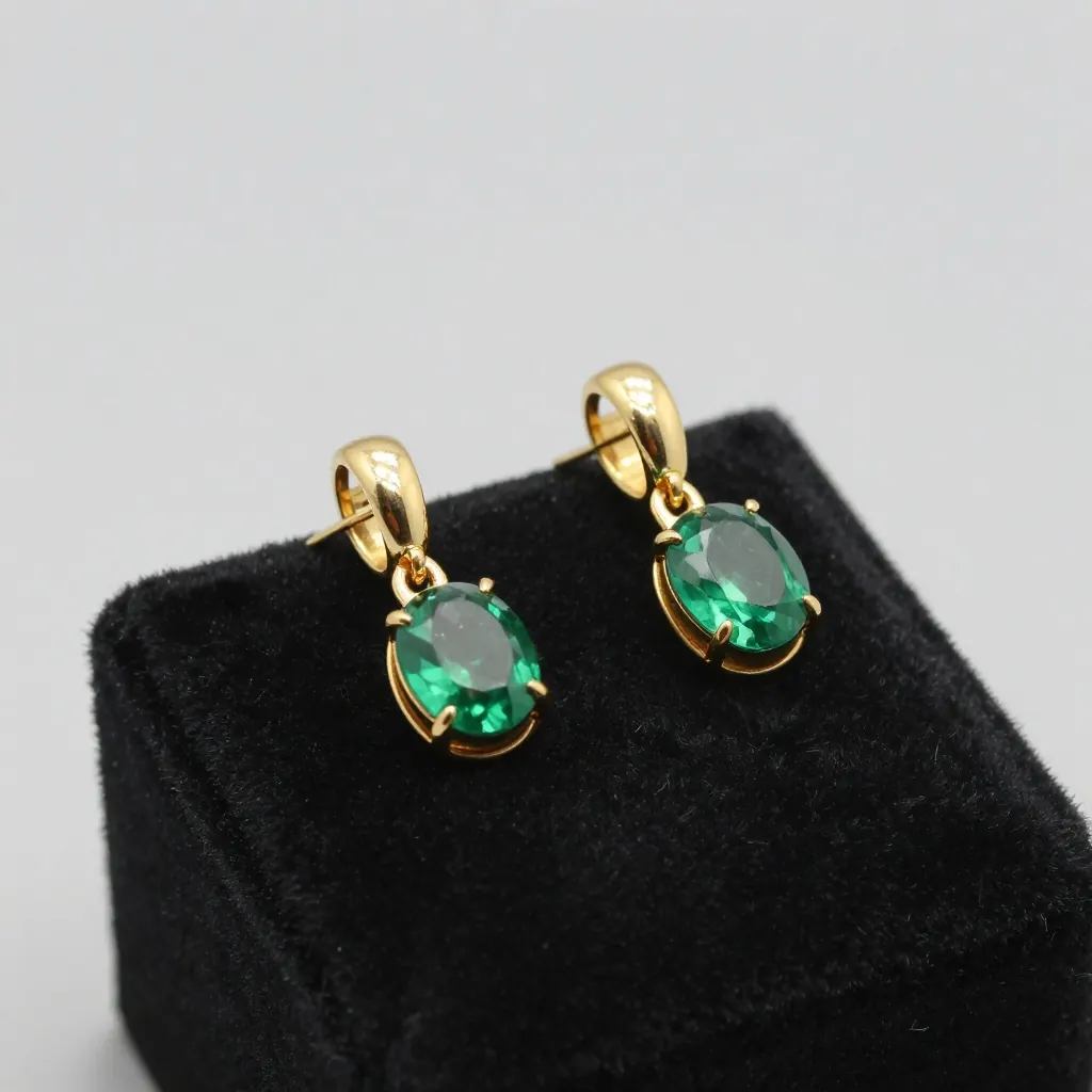Crisp product photograph of a pair of polished gold earrings with emerald stones on a black velvet pedestal, single soft spotlight, sharp reflections, luxury jewelry photography, no text, no logo